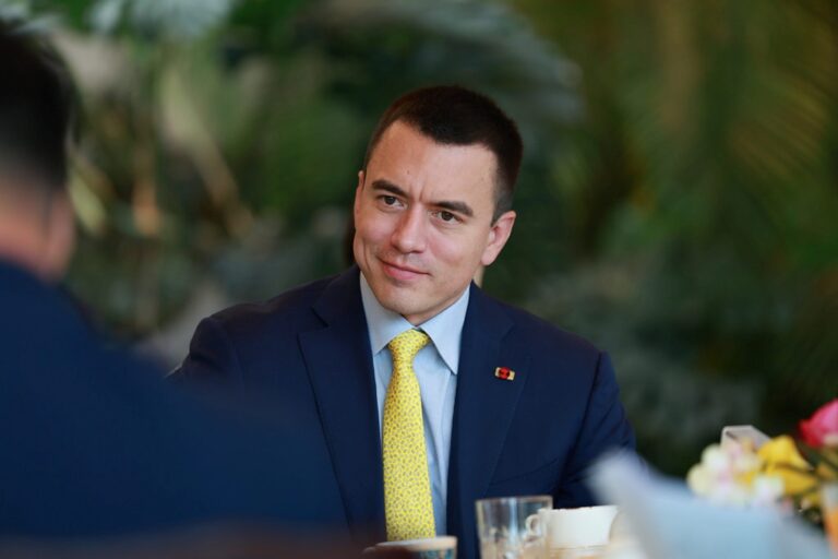 Daniel Noboa, con traje azul marino y corbata amarilla, participa en una reunión formal con fondo desenfocado de vegetación.