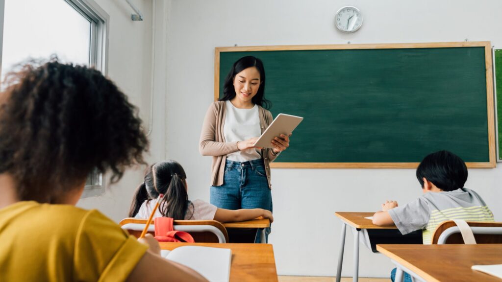 Maestra en Japón utilizando una tableta frente a sus alumnos en el salón de clases, reflejando las extensas jornadas laborales que enfrentan los docentes Japón.