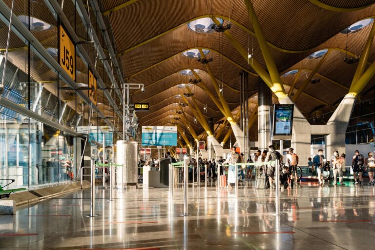 Interior iluminado de la terminal de salidas del aeropuerto Adolfo Suárez Madrid-Barajas, con viajeros en fila y señales de puertas J58 y J59 visibles.