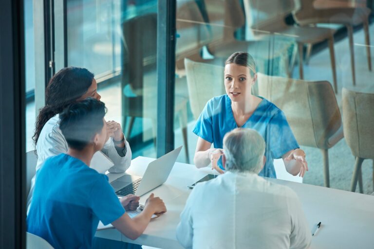 Grupo de profesionales del sector salud reunidos en una mesa de trabajo, dialogando sobre estrategias de capacitación y asignación de plazas, en un entorno institucional con ventanales de vidrio.