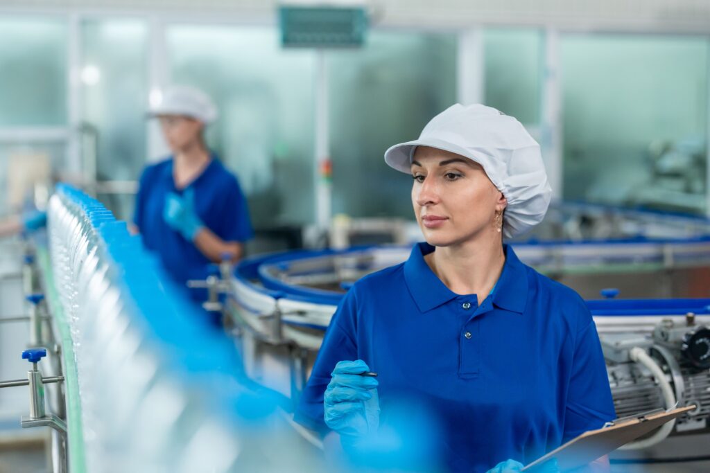 Mujer con uniforme azul y gorro de seguridad supervisando la línea de embotellado en una fábrica, símbolo del liderazgo femenino en entornos industriales y sindicales.