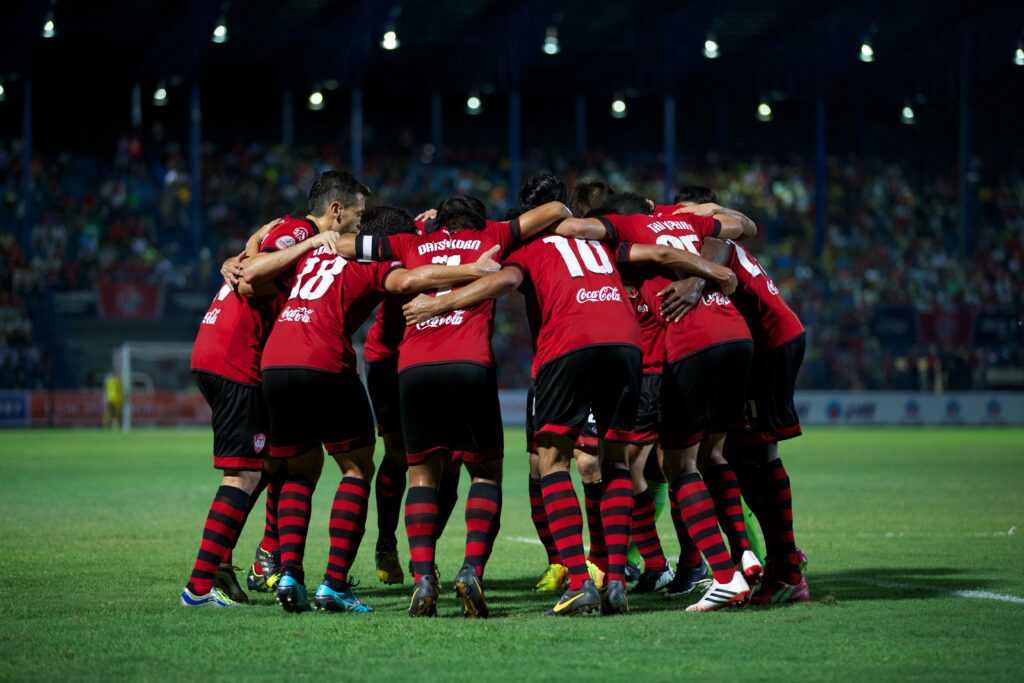 Jugadores de fútbol con uniforme rojo y negro en círculo abrazados, mostrando unidad y trabajo en equipo antes del inicio del partido.