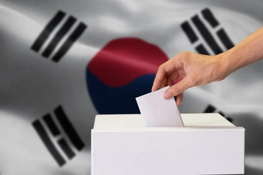 Mano introduciendo papeleta en urna durante votación en Corea del Sur, con la bandera nacional de fondo, representando la aprobación de la reforma laboral en 2025.