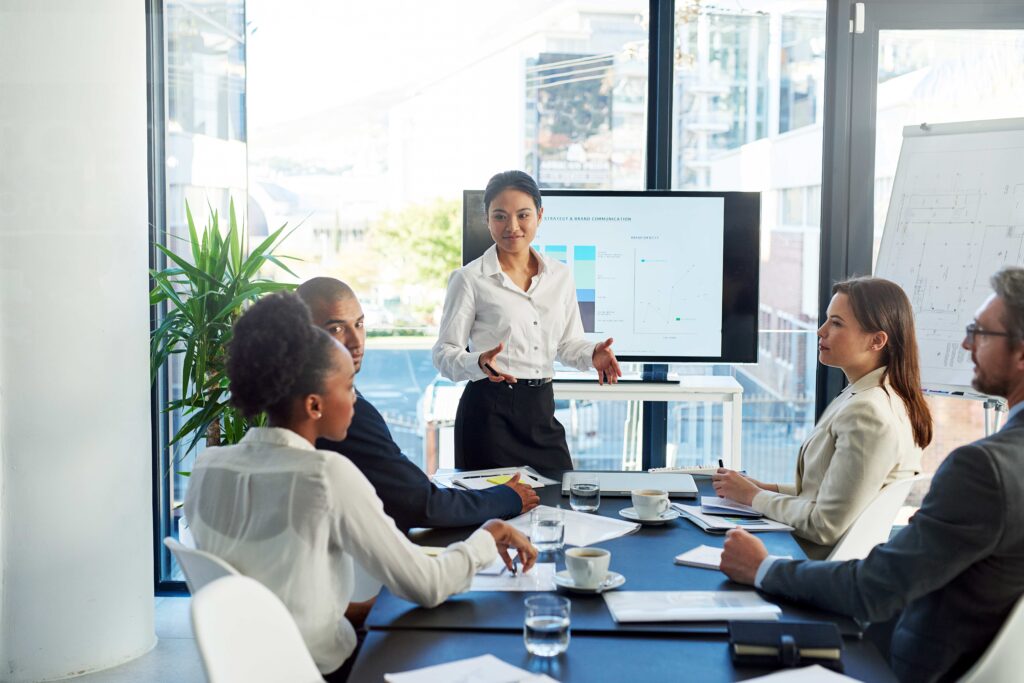 Grupo de profesionales en junta laboral discutiendo estrategias sobre el regreso a la oficina, con presentación en pantalla y liderazgo femenino al frente