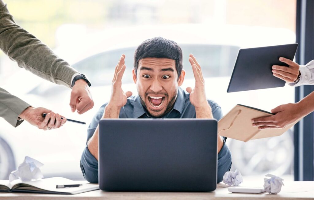 Hombre estresado frente a computadora rodeado de tareas y presión, ejemplo de riesgos laborales que la salud laboral busca prevenir.